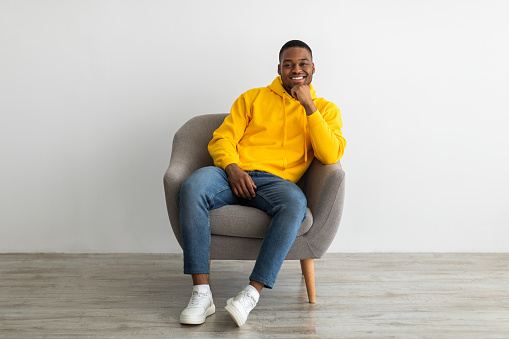 Successful Black Millennial Guy Smiling To Camera Touching Chin Sitting In Armchair Near Gray Wall Indoors, Wearing Yellow Hoodie. African American Man Relaxing Posing At Home. Full Length