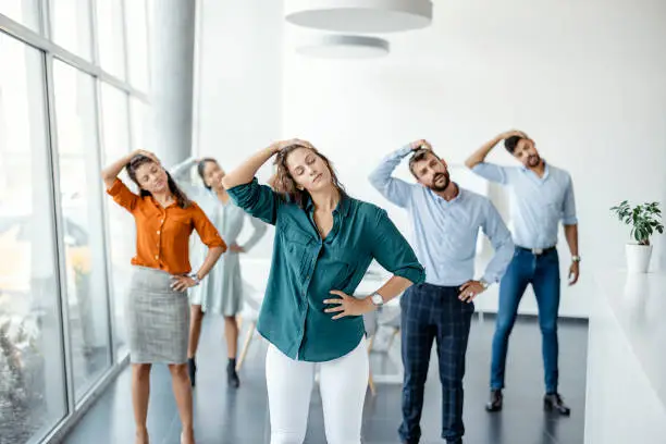 Diverse colleagues doing neck stretching exercise standing in office. Multiethnic young office employees do workout exercises during break at work