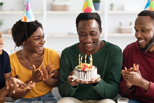 Handsome black birthday guy holding cake with lit candles and smiling, making wish before blowing out. Cheerful african american millennial friends making surprise for young man, closeup photo