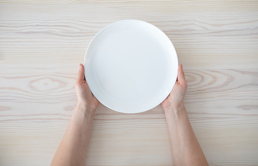 Woman holding empty plate waiting for food, sitting on wooden table, top view, copy space. Hunger and lunch time concept