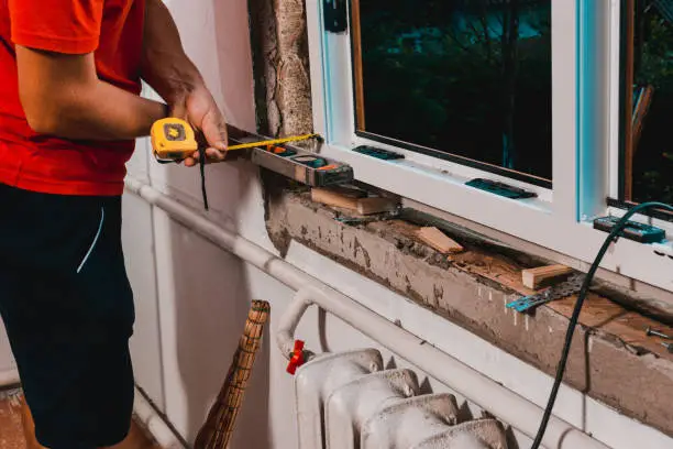 The man performs installation work with the window, for this he also uses a tape measure, measures distances and fastens the window to the wall.2020