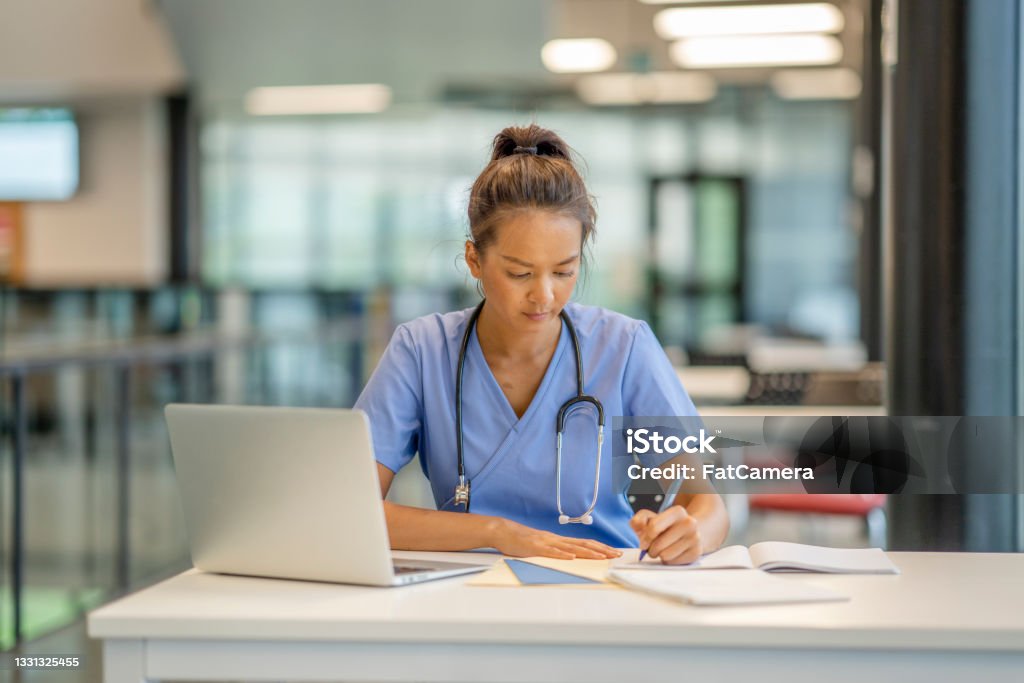 Medical student studying A woman wearing scrubs diligently studies at a desk in a modern educational building. Nurse Stock Photo