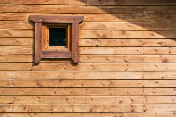The wall of a wooden house is sheathed with brown horizontal planks. There is a small square window with a casing and an oblique roof shade. Background.