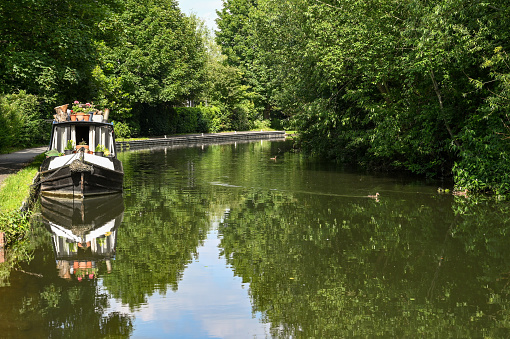Newbury, Berkshire, England - June 2021: Front of a houseboat moored in a marina on the Kennet and Avon Canal near the centre of Newbury, with reflection in still water.