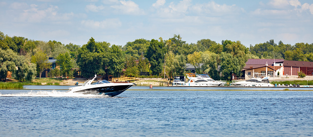 The motor boat moves quickly along the water surface of the river. In the background, the coastline in the summer with a green park, a pier and residential private house. Copy space.