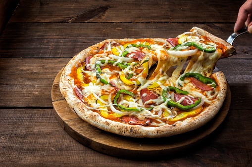 Pizza with vegetables (yellow and green bell pepper, onion) and pepperoni. A hand with a utensil is lifting a slice with appetizingly melted cheese.