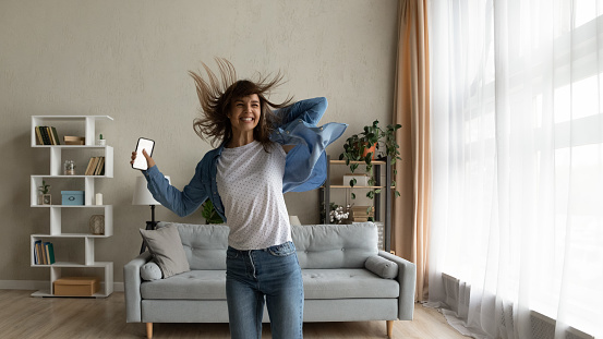 Cheery woman feels overjoyed jumps dance in living room hold smart phone with mock up white screen. Female celebrate unbelievable great news received. Fun, moment of victory, candid emotions concept