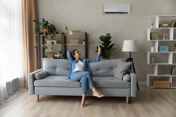Happy young woman relax on couch in living room holding remote controller turn on air conditioner set comfort temperature, enjoy fresh air at summer day inside modern flat, ac, climate control concept
