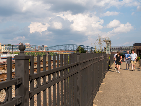 Pittsburgh, Pennsylvania, USA July 25, 2021 People walking along a walkway at Station Square with a wrought iron fence next to train tracks and the Smithfield Street bridge in the background