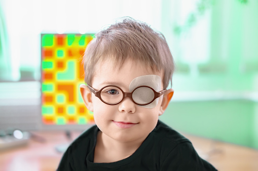 A little boy wearing glasses and an eye patch (plaster, occluder) undergoes a hardware vision treatment to prevent amblyopia and strabismus (squint, lazy eye). Child congenital vision disease problem.