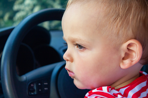 Small caucasian baby boy as driver. Sitting in driver's seat in car. Travel with kids concept. Baby looking at something through the open car window.