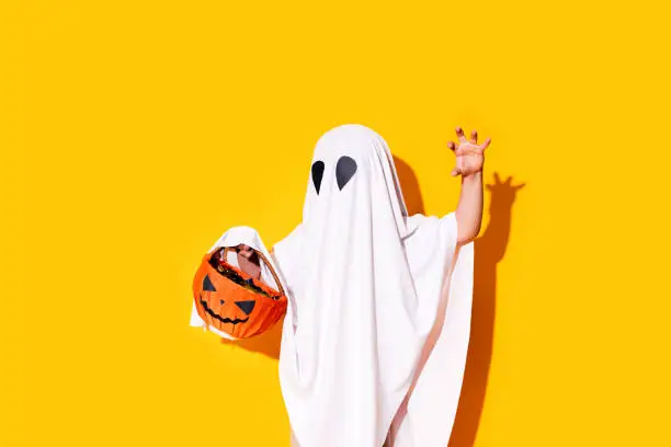 Child in ghost costume holding pumpkin basket.