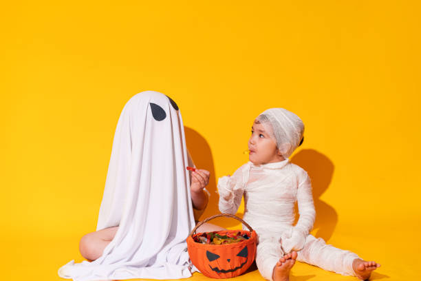 ハロウィーンのためのマミアと幽霊の衣装を着た子供たちは、黄色の背景に甘いもののバスケットの前に座っています - vegies vegetable basket residential structure ストックフォトと画像