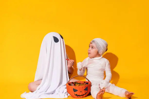 Photo of Children in mamia and ghost costumes for halloween sit in front of basket of sweets on yellow background