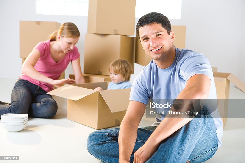 Family between boxes Young parents and their daughter sitting beside cardboard boxes. Young girl sitting in box. They're smiling. Focused on man who sitting in front. 2-3 Years Stock Photo