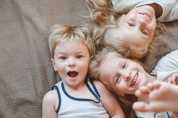침대에 누워 있는 세 아이의 초상화. 카메라를 보면서 웃고 있는 자매와 형제 - child laughing blond hair three people 뉴스 사진 이미지