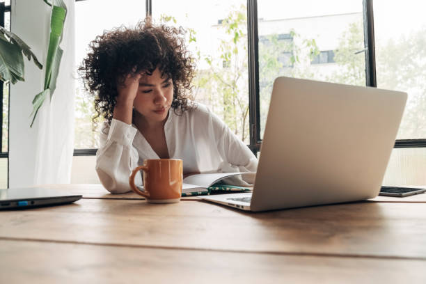 joven afroamericana cansada, agotada de trabajar estudiando duro. aburrido y frustrado mirando la computadora portátil. cabeza descansando sobre la mano. espacio luminoso grandes ventanales. - frustración fotografías e imágenes de stock