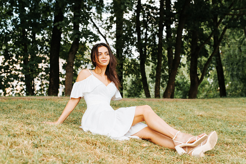 Young brunette woman in white dress chilling in the park, lying on green grass of the lawn, resting and enjoying the freedom and leisure of a summer day. Beautiful girl in nature looking to the side.