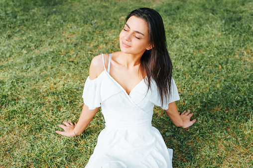 Charming young brunette woman in white light dress having a rest with closed eyes on green grass lawn in park outdoors, high angle view. Beautiful mixed race lady enjoying freedom, lifestyle outside.