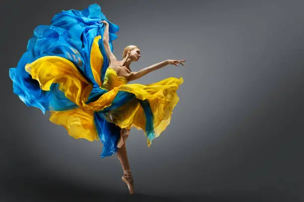 Beautiful Woman Ballet Dancer Jumping in Air in Colorful Fluttering Dress. Graceful Ballerina Dancing in Yellow Blue Gown over Gray Studio Background. Dynamic Motion Dance