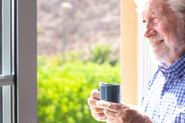 istock Smiling senior man at the window holding a coffee cup looking out 1331431181