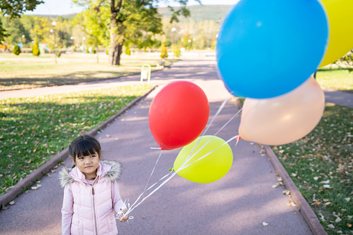 A small Asian girl wearing a winter coat is holding a bunch of five colorful balloons, standing on a footpath in a public park on a bright sunny day