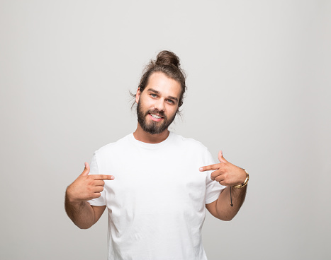 Portrait of cheerful bearded young man with hair bun wearing white t-shirt. Male student pointing with index fingers at t-shirt. Studio shot, grey background.