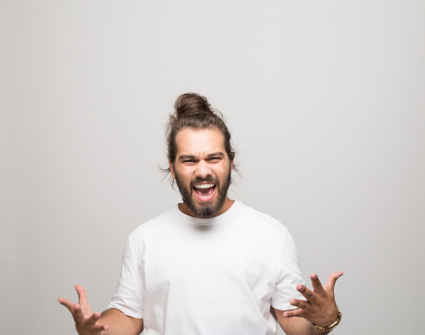Portrait of happy bearded young man with hair bun wearing white t-shirt. Male student shouting at camera. Studio shot, grey background.