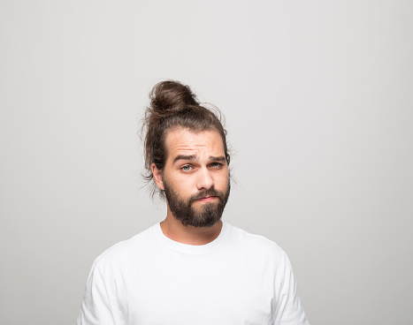 Portrait of bearded young man with hair bun wearing white t-shirt. Confused male student looking at camera. Studio shot, grey background.