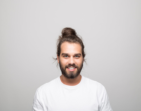 Portrait of cheerful bearded young man with hair bun wearing white t-shirt. Male student laughing at camera. Studio shot, grey background.