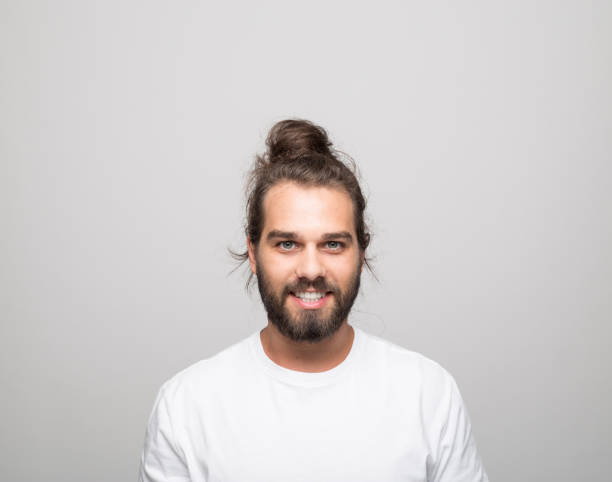 Headshot of happy young man Portrait of cheerful bearded young man with hair bun wearing white t-shirt. Male student laughing at camera. Studio shot, grey background. long hair stock pictures, royalty-free photos & images