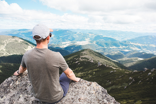 young strong man sitting on the rock at the top of the mountains peak enjoying the view. hiking concept