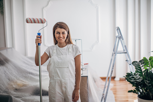 One woman, portrait of a beautiful female holding a paint roller in living room at home. She is painting walls.