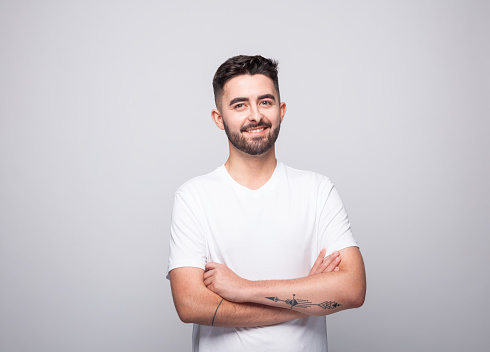 Portrait of cheerful bearded young man wearing white t-shirt. Male student standing with arms crossed and smiling at camera. Studio shot, grey background.