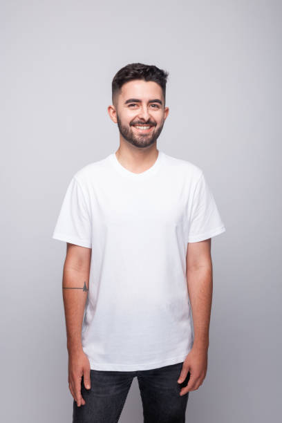 Portrait of cheerful bearded young man wearing white t-shirt. Male student smiling at camera. Studio shot, grey background.