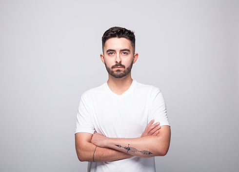Portrait of handsome bearded young man wearing white t-shirt. Male student standing with arms crossed and looking at camera. Studio shot, grey background.