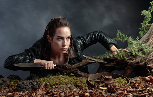 Portrait of a young woman with a knife. A serious girl in a black leather jacket with a knife climbs out of the pit in the forest. Copy space.