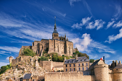 Normandy, France, August 29, 2019: Looking at the huge city walls which surround Mont Saint-Michel mirrored on small puddles, while low tide flush out the water around the Mont Saint-Michel island. Le Mont-Saint-Michel or in english Saint Michael's Mount is a tidal island and mainland commune in Normandy, France. The island lies approximately one kilometre (0.6 miles) off the country's north-western coast, at the mouth of the Couesnon River near Avranches and is 7 hectares (17 acres) in area. The mainland part of the commune is 393 hectares (971 acres) in area so that the total surface of the commune is 400 hectares (988 acres). As of 2017, the island has a population of 30. \nAs of 2017, the island has a population of 30. Mont-Saint-Michel and its bay are on the UNESCO list of World Heritage Sites. It is visited by more than 3 million people each year. Over 60 buildings within the commune are protected in France as monuments historiques.