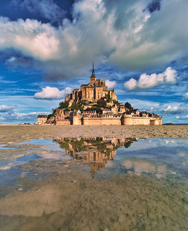 Normandy, France, August 29, 2019: Mont Saint-Michel mirrored on small puddles, while low tide flush out the water around the Mont Saint-Michel island. Le Mont-Saint-Michel or in english Saint Michael's Mount is a tidal island and mainland commune in Normandy, France. The island lies approximately one kilometre (0.6 miles) off the country's north-western coast, at the mouth of the Couesnon River near Avranches and is 7 hectares (17 acres) in area. The mainland part of the commune is 393 hectares (971 acres) in area so that the total surface of the commune is 400 hectares (988 acres). As of 2017, the island has a population of 30. \nAs of 2017, the island has a population of 30. Mont-Saint-Michel and its bay are on the UNESCO list of World Heritage Sites. It is visited by more than 3 million people each year. Over 60 buildings within the commune are protected in France as monuments historiques.