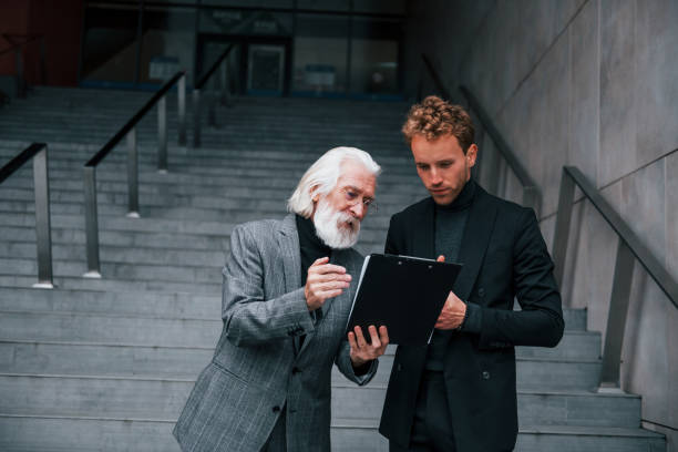 istock Holding notepad. Young guy with senior man in elegant clothes is outdoors together. Conception of business 1331446585