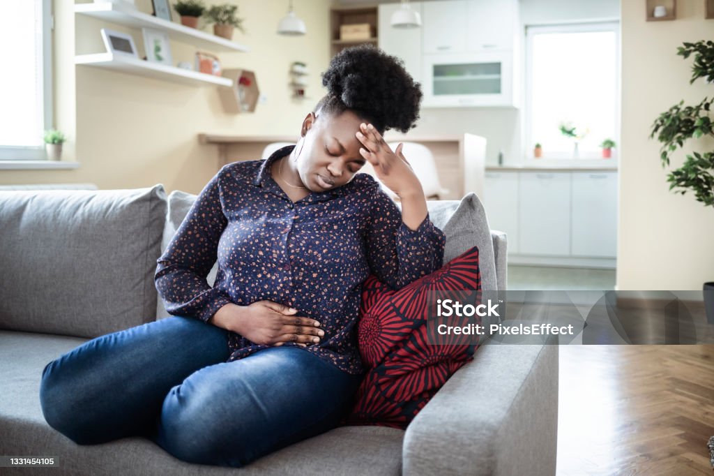 The pain is worsening by the minute Young sick african american plus size woman with hands holding pressing her crotch lower abdomen. Medical or gynecological problems, healthcare concept. Young woman suffering from abdominal pain while sitting on sofa at home Abdomen Stock Photo