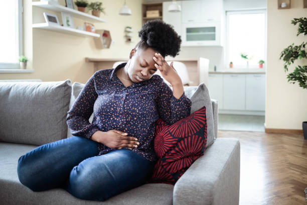 The pain is worsening by the minute Young sick african american plus size woman with hands holding pressing her crotch lower abdomen. Medical or gynecological problems, healthcare concept. Young woman suffering from abdominal pain while sitting on sofa at home stomach stock pictures, royalty-free photos & images