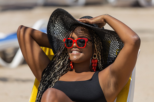 Close-up of an African-American woman on the beach lying on a deck chair wearing a black hat and sunglasses. Cheerful adult woman enjoying a summer day at the beach.