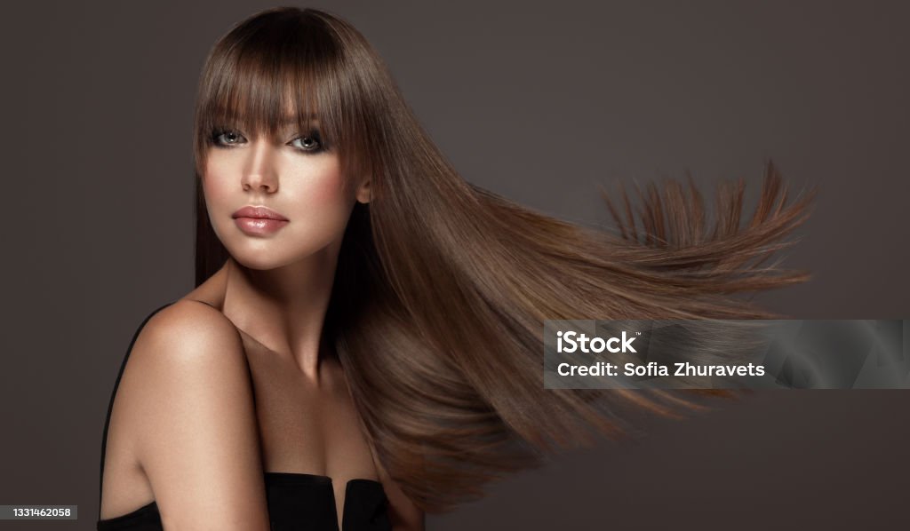 Young, brown haired beautiful model with long, straight, well groomed hair. Hair care and hairdressing art. Attractive young woman with chubby, well shaped lips and green eyes with smoky eyeshadows makeup is demonstrating long, straight, dense hairstyle with long, straight fringe.Flying hair. Straight Hair Stock Photo