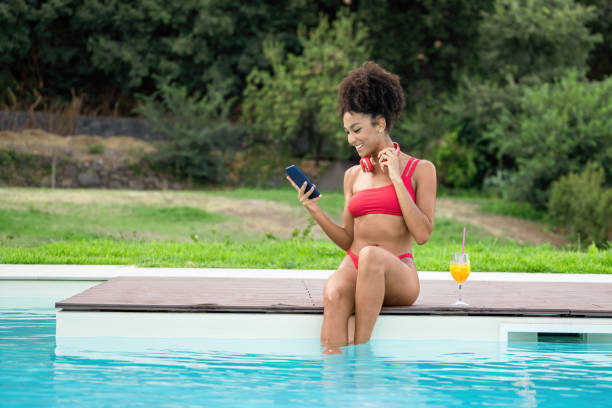 afroamerykańska dziewczyna siedząca na skraju basenu uśmiecha się podczas rozmów wideo ze smartfonem, trzymając szklankę soku owocowego. - tourist resort audio zdjęcia i obrazy z banku zdjęć
