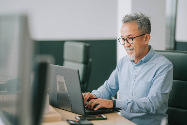 uomo anziano cinese asiatico con i peli del viso usando la digitazione del laptop lavorando in ufficio open plan - gray hair immagine foto e immagini stock