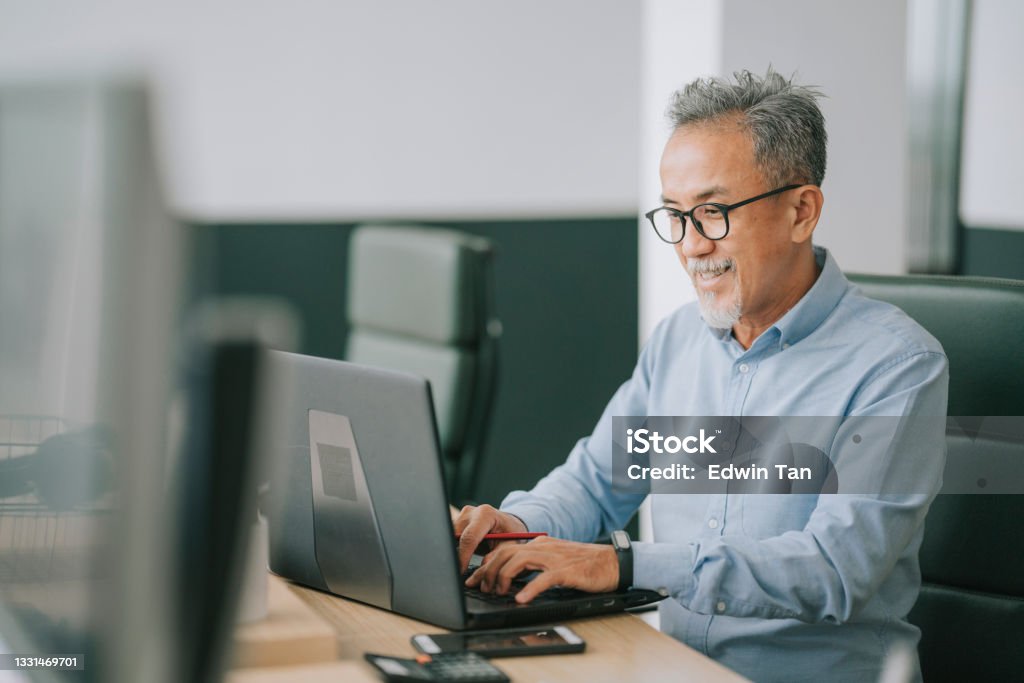 Asian chinese senior man with facial hair using laptop typing working in office open plan Office Stock Photo