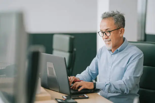 Photo of Asian chinese senior man with facial hair using laptop typing working in office open plan