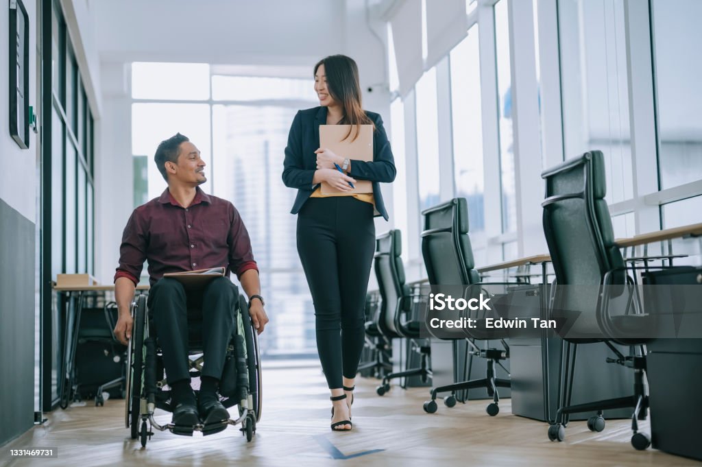 Indian white collar male worker in wheelchair having cheerful discussion conversation with his female asain chinese colleague coworking at walkway corridor Multiracial Group Stock Photo