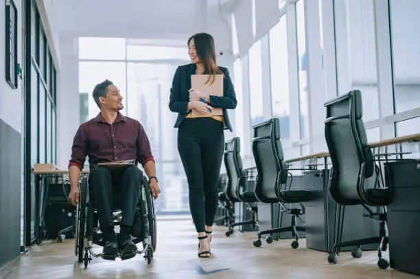 Photo of Indian white collar male worker in wheelchair having cheerful discussion conversation with his female asain chinese colleague coworking at walkway corridor